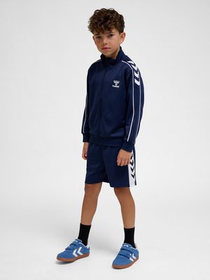 Junge steht und trägt eine marineblaue Trainingsjacke und Shorts mit weißen Streifen, schwarze Socken und blaue Turnschuhe vor einem schlichten grauen Hintergrund.