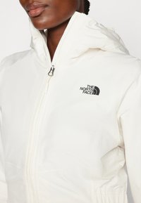 Giacca impermeabile bianca con cappuccio, caratterizzata da un logo nero e dettagli con cerniera. Texture liscia e design aderente.