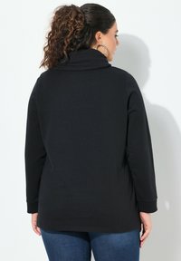 Sudadera negra de manga larga con cuello estilo cowl, fabricada en un material suave, corte relajado y dobladillo recto, combinada con vaqueros azules.