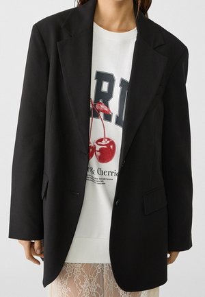 Frau trägt schwarzen Blazer über weißem Sweatshirt mit großem Kirschmotiv und Spitzenrock.