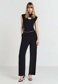 Top senza maniche nero con accento a forma di chiave, abbinato a pantaloni neri a vita alta e plissettati e sandali neri con tacco, creando una silhouette elegante.