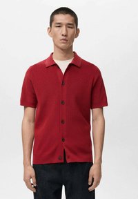 Maglia rossa a maniche corte con collo, caratterizzata da una chiusura frontale con bottoni e tessuto strutturato. Design semplice, abbinata a denim scuro.