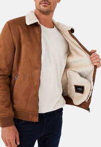 Brown suede jacket with a cream shearling collar and interior. Features a zipper closure and side pocket with a fabric label, worn over a light shirt.