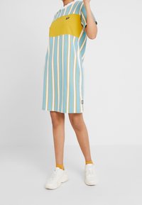 Randig klänning med en gul blockpanel, som har blå och vita vertikala ränder samt en liten logotyp. Tillverkad av mjuk, lättviktig tyg.