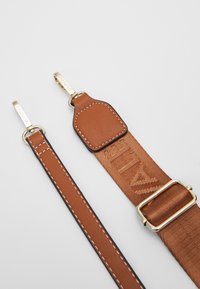 Brun justerbar rem gjord av vävt tyg och läderdetaljer; har guldtonade metalldetaljer och präglad logotyp. Slät textur med kontrasterande sömmar.