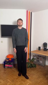 Grå strikkegenser, kort modell, kombinert med svarte vidbukser. Mørkebrune skinnsko. Innendørs setting med dekorative elementer.