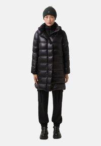 Czarna kurtka puchowa o błyszczącej powierzchni, z kapturem, zamkiem błyskawicznym i pikowanym wzorem. Połączona z czarnymi spodniami i masywnymi butami.