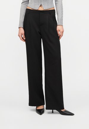 Kobieta ubrana w czarne spodnie o szerokich nogawkach, szary skrócony kardigan i czarne czółenka na niskim obcasie ze szpiczastym noskiem, stoi na białym tle.