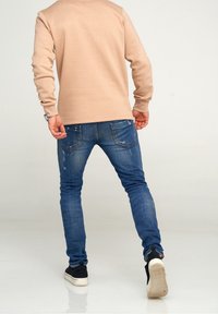 Beige sweatshirt med långa ärmar, i kombination med tajta blå jeans med slitna detaljer och svarta sneakers med vita sulor.