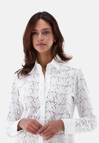 Weißes Broderie Anglaise Hemd mit kragenfreiem Ausschnitt, Knopfverschluss und blumigen Ausschnitten. Lange Ärmel mit weißen Manschetten.
