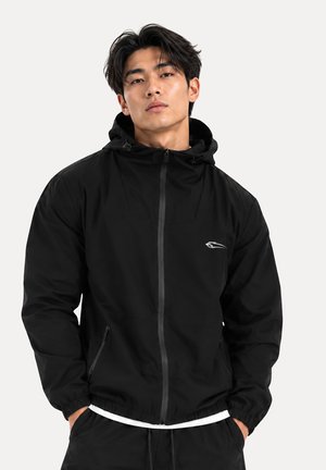 Jeune homme portant une veste à capuche noire zippée, les mains dans les poches, sur un fond clair uni.