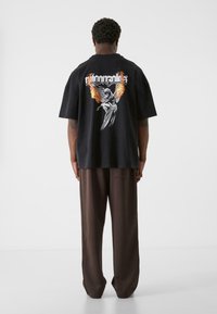 Sort oversized t-shirt med grafisk design med flammer og en vinget figur på ryggen, parret med løse brune bukser.