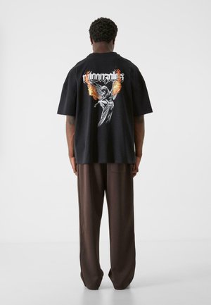 Svart oversized t-skjorte med grafisk design av flammer og en bevinget figur på ryggen, sammen med løse brune bukser.