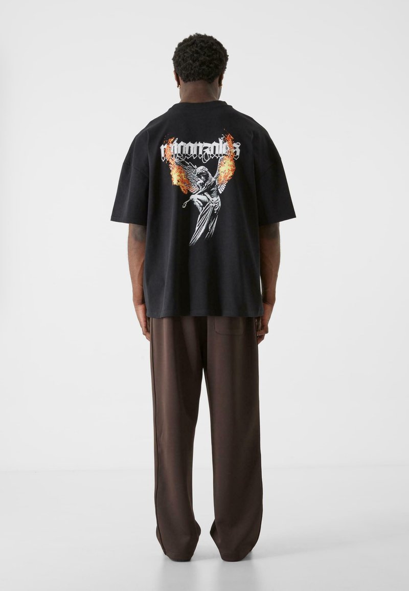 Svart oversized t-skjorte med grafisk design av flammer og en bevinget figur på ryggen, sammen med løse brune bukser.