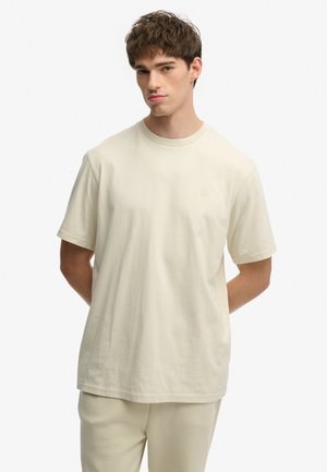 Ung mand med kort brunt hår iført en ensfarvet beige oversized T-shirt og matchende bukser, stående med hænderne på ryggen.