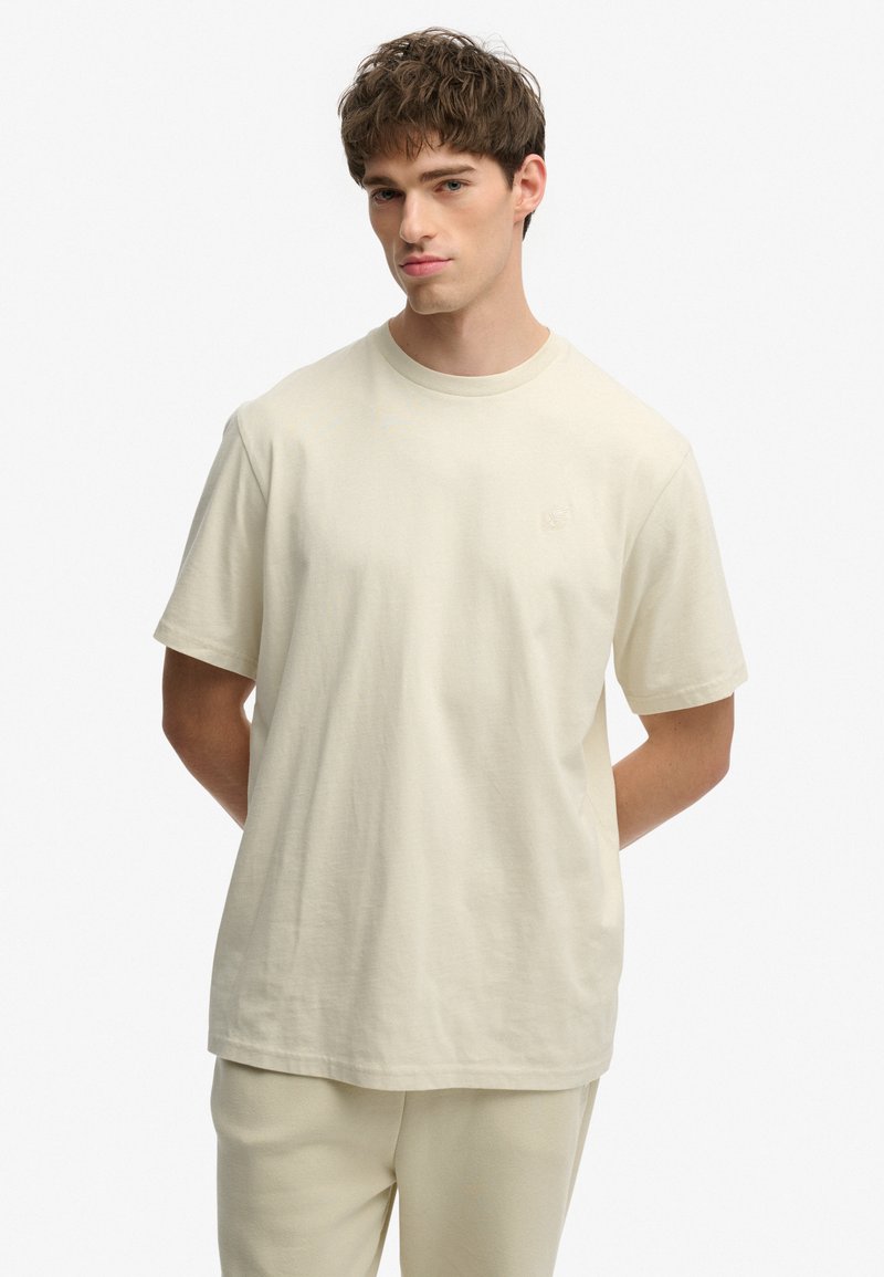 Jeune homme aux cheveux courts et bruns portant un t-shirt beige uni et oversize ainsi qu’un pantalon assorti, debout les mains dans le dos.