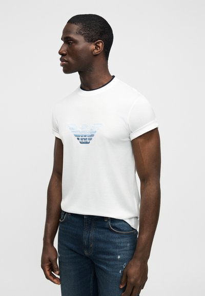 Λευκό βαμβακερό μπλουζάκι με κοντά ρολαριστά μανίκια και μπλε βαθμιαίο λογότυπο στο στήθος. Συνδυασμένο με σκούρο μπλε τζιν παντελόνι.