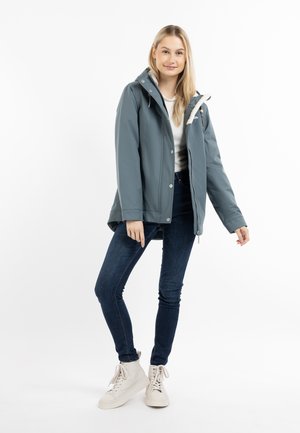 Blå vandtæt jakke med hætte, frontlynlås og trykknapper. Bæret over en lys sweater og kombineret med mørke jeans og hvide sneakers.