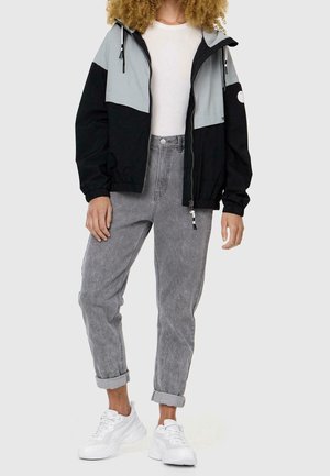Person iført en sort og grå lynlåstrøje, hvid skjorte, opkørte grå jeans og hvide sneakers, stående mod en ensfarvet baggrund.