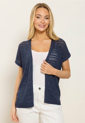 Donna sorridente che indossa un cardigan a maniche corte a maglia aperta blu navy sopra una canotta bianca e pantaloni bianchi, in piedi contro uno sfondo semplice.