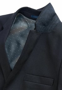 Blazer bleu marine foncé avec un col texturé élégant. La doublure intérieure présente un tissu gris avec des petits pois bleu clair. Poche unique à l'avant.