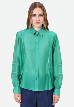 Femme portant une chemise verte en soie à manches longues avec des coutures décoratives et un pantalon bleu foncé, debout devant un fond uni.