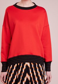 Sudadera roja con cuello y puños de canalé negros, con acentos de costura dorada. El corte es relajado y tiene un acabado texturizado.