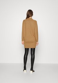 Svetr dress v barvě velbloudí, ke kolenům, s dlouhými rukávy a žebrovanými manžetami, nošený přes černé, přiléhavé kožené legíny a kotníkové boty na podpatku.