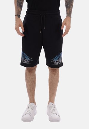 Mand iført sorte shorts med snøre samt blå og grå fjerdesign nær lommekant, hvide sneakers og en sort skjorte, står med armene afslappet.