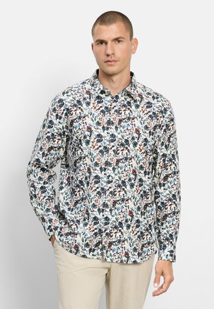 Mann trägt ein langärmeliges weißes Hemd mit blauen, braunen und grünen Blumenmustern, kombiniert mit hellbeigen Hosen, steht vor weißem Hintergrund.