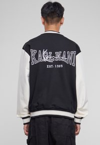 Veste bomber noire et crème avec le lettrage "KARL KANI" au dos, des poignets et un ourlet côtelés, et une texture douce.