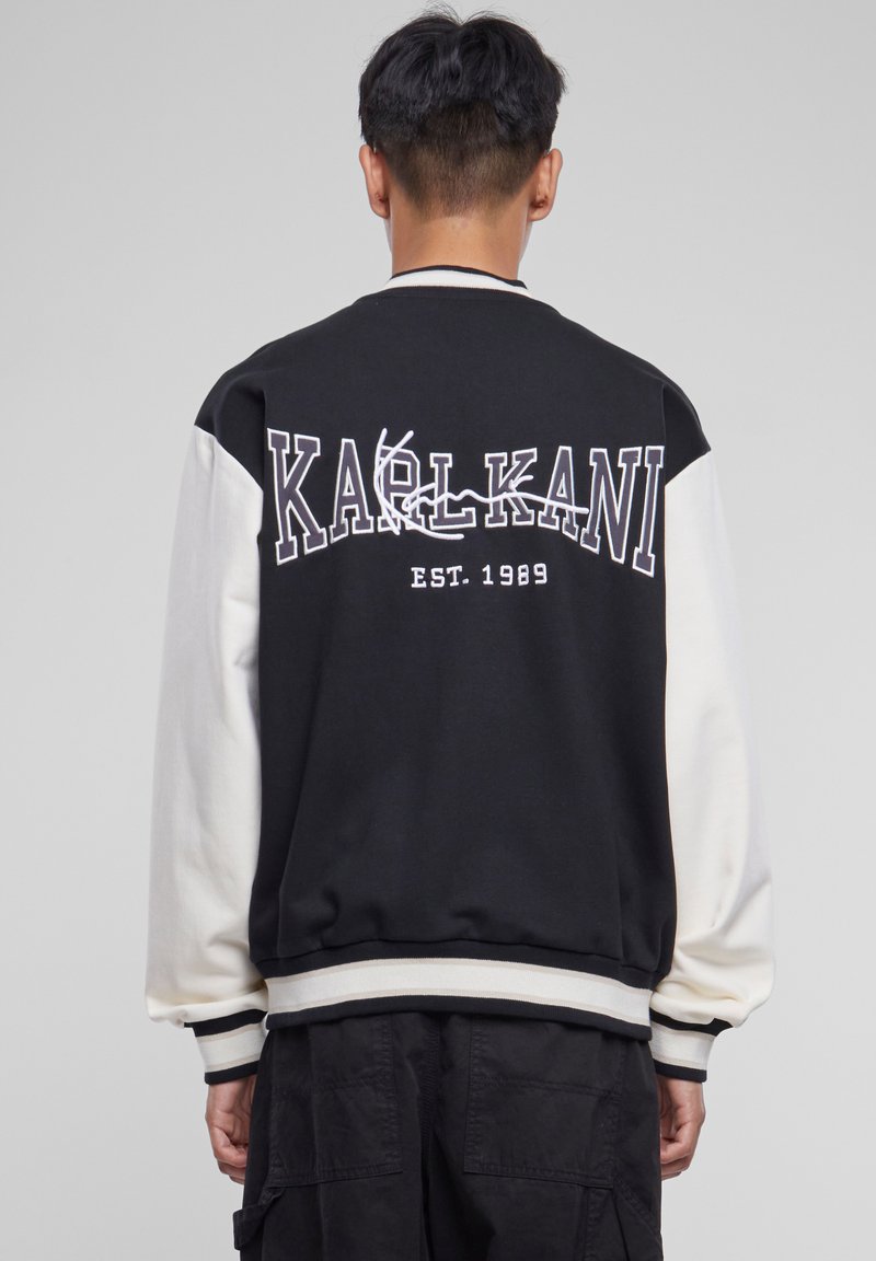 Veste bomber noire et crème avec le lettrage "KARL KANI" au dos, des poignets et un ourlet côtelés, et une texture douce.