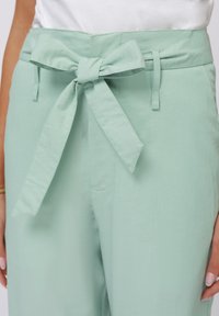 Mint green trousers with a wide waistband featuring a tied bow detail. Made of a soft, lightweight fabric with a smooth texture.