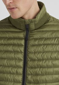 Olivgrüne Daunenjacke mit hohem Kragen, ausgestattet mit horizontalen Nähten und einem schwarzen Reißverschluss. Strukturelle Akzente im Bereich des Kragens.