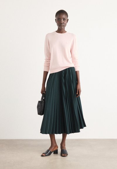 Pull rose clair, jupe plissée verte foncée, sandales à talons noirs, et un sac à main tissé noir. Texture lisse et lignes épurées.