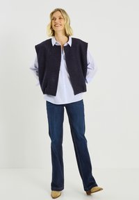 Chaleco sin mangas texturizado azul marino sobre una camisa de rayas azul claro, combinado con vaqueros acampanados oscuros y calzado beige.