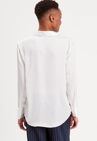 Chemise blanche à manches longues avec une texture lisse et brillante, dotée d'un col en V, de poignets boutonnés et d'un ourlet arrondi à l'arrière.