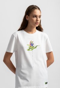 Witte katoenen t-shirt met een kleurrafisch van een hand en groene bladeren op de voorkant, ronde halslijn en korte mouwen.