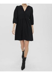 Robe noire à manches bouffantes avec un décolleté en V, une patte de boutonnage à l'avant, un corsage ajusté et une jupe évasée, réalisée dans un tissu lisse. Portée avec des bottines noires.