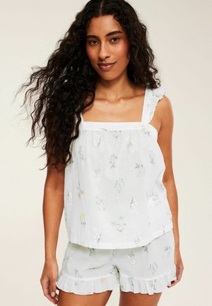 Femme aux longs cheveux bouclés portant un haut blanc fleuri sans manches et un short à volants assorti sur un fond uni.