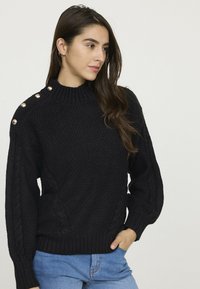 Pull en maille noir avec un col montant, des accents sur l'épaule droite ornés de boutons dorés, et des motifs de tricot en relief sur les manches.