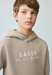 Bež dukserica od glatkog materijala s velikim prednjim printom "BAKER BY TED BAKER." Materijal je mekan s opuštenim krojem i prednjim džepom.