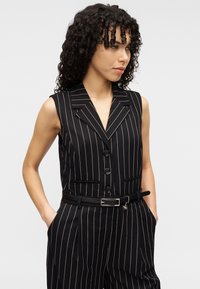 Combinaison sans manches à rayures fines noires avec un col en V, des boutons sur le devant et des poches latérales. Comprend une ceinture noire avec une boucle argentée.