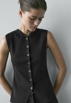 Vrouw met achtergebonden haar die een mouwloos zwart vest met knopen draagt, kijkt neerwaarts tegen een effen lichtgrijze achtergrond.