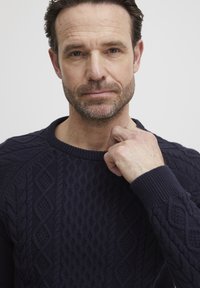 Pull en tricot bleu marine avec des motifs en relief, un col rond et des poignets côtelés. Gros plan sur une main au niveau du col.