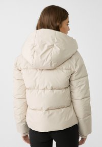 Ljusbeige pufferjacka med huva, med ett quiltat mönster och elastiska ärmslut. Slät textur och kort silhuett.