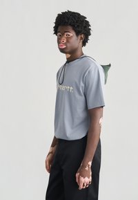 Γκρι βαμβακερό t-shirt με κοντά μανίκια και λογότυπο της μάρκας, σε συνδυασμό με μαύρο παντελόνι. Διαθέτει ένα πράσινο αξεσουάρ που κρέμεται από τον ώμο.