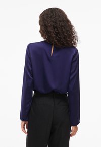 Kvinna med lockigt hår iförd en långärmad lila blus och svarta byxor, sedd bakifrån mot en vit bakgrund.