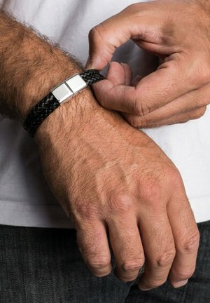 Mand fastgør sort flettet læderarmbånd med sølvspænde på venstre håndled, iført hvid skjorte og mørke bukser.