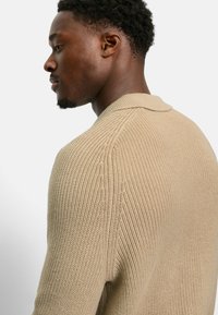 Beige gestrickter Pullover mit einem strukturierten Muster, einem gerippten Kragen und eingesetzten Ärmeln. Das Kleidungsstück hat eine lässige Passform und einen weichen Stoff.